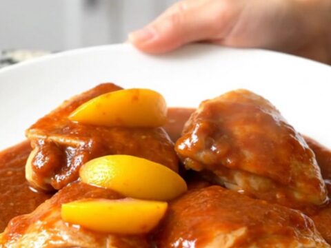 A close-up of tender chicken thighs coated in a rich, reddish-orange gochujang peach sauce in a slow cooker.