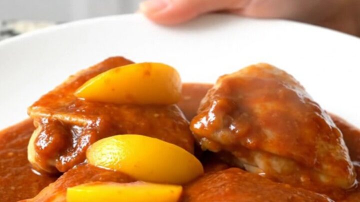 A close-up of tender chicken thighs coated in a rich, reddish-orange gochujang peach sauce in a slow cooker.