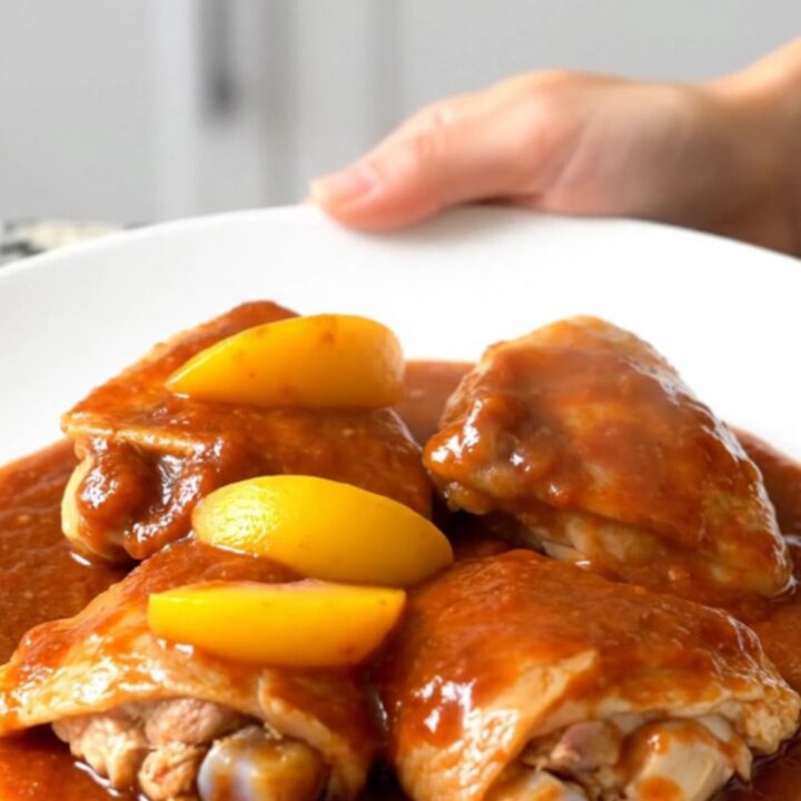 A close-up of tender chicken thighs coated in a rich, reddish-orange gochujang peach sauce in a slow cooker.