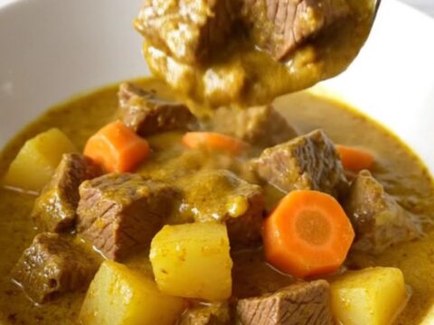 A close-up of a slow cooker filled with beef, potatoes, and carrots in a rich, golden coconut curry sauce.