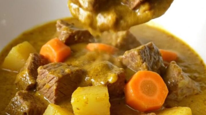 A close-up of a slow cooker filled with beef, potatoes, and carrots in a rich, golden coconut curry sauce.