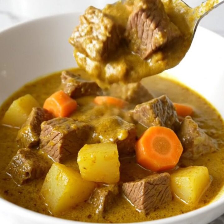 A close-up of a slow cooker filled with beef, potatoes, and carrots in a rich, golden coconut curry sauce.