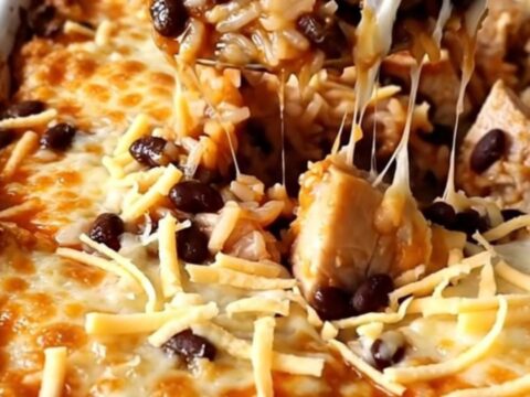 A top-down view of a rectangular baking dish filled with raw rice, chicken pieces, red enchilada sauce, and black beans.