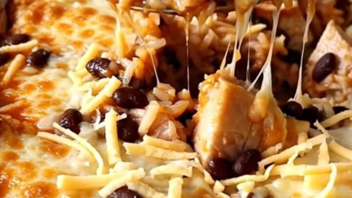 A top-down view of a rectangular baking dish filled with raw rice, chicken pieces, red enchilada sauce, and black beans.