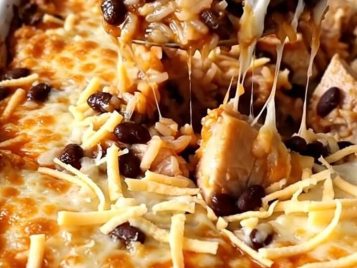 A top-down view of a rectangular baking dish filled with raw rice, chicken pieces, red enchilada sauce, and black beans.