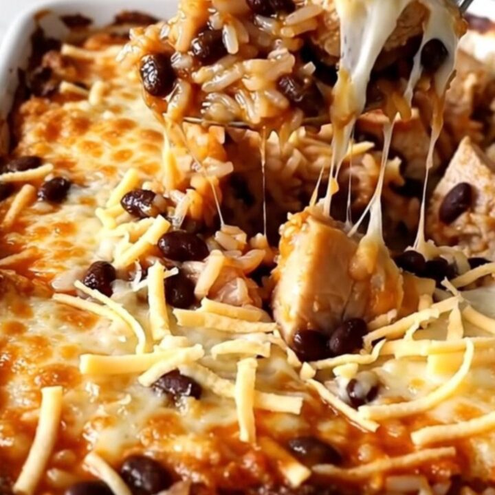 A top-down view of a rectangular baking dish filled with raw rice, chicken pieces, red enchilada sauce, and black beans.