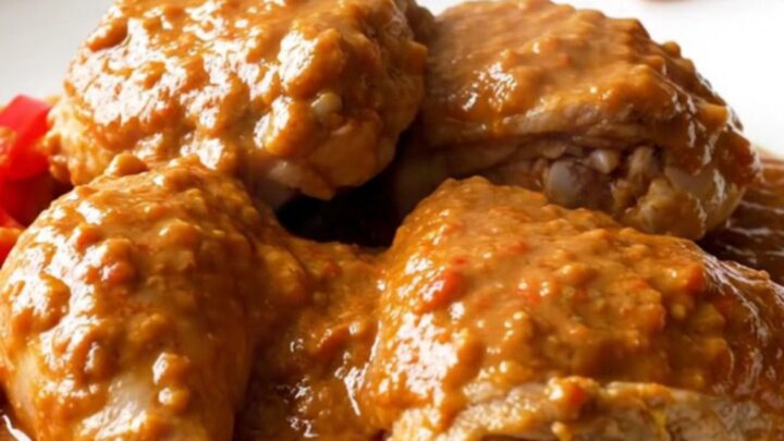 A close-up of tender chicken thighs covered in a thick, creamy peanut tomato sauce in a white slow cooker.
