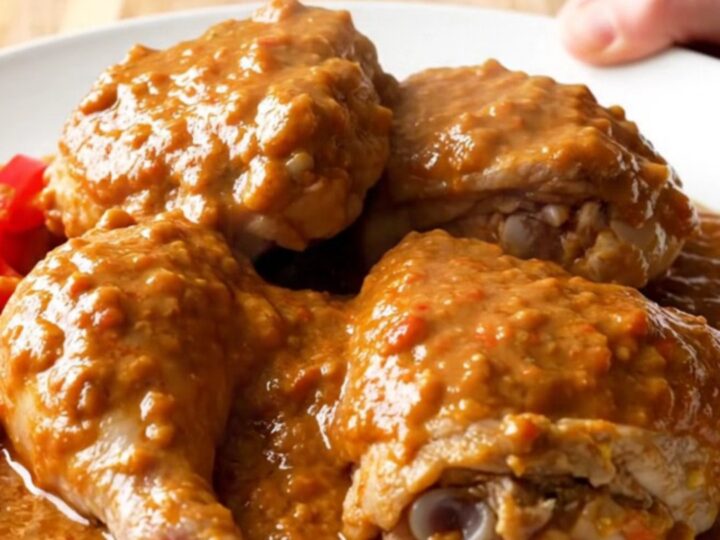 A close-up of tender chicken thighs covered in a thick, creamy peanut tomato sauce in a white slow cooker.