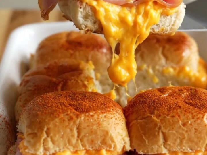 A close-up of golden-brown sliders in a white ceramic dish, with visible melted pimento cheese and ham.