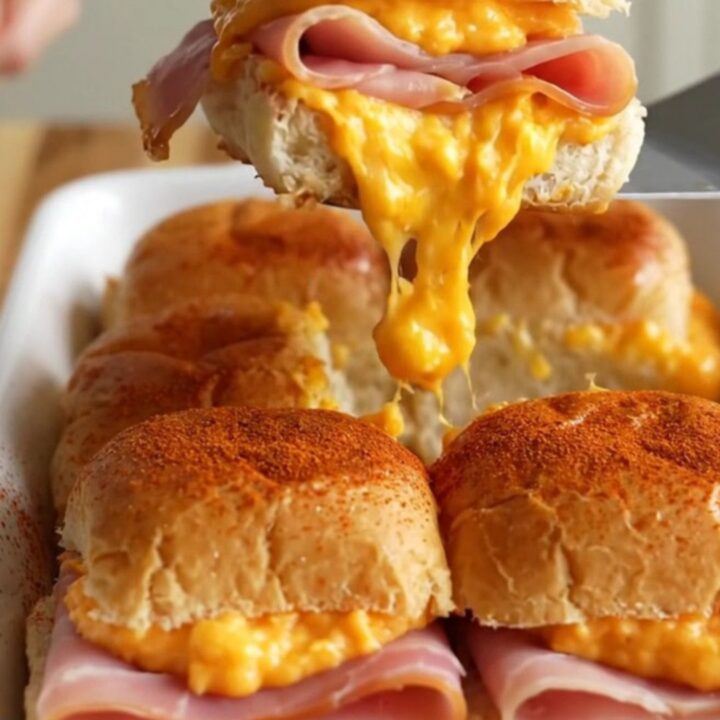 A close-up of golden-brown sliders in a white ceramic dish, with visible melted pimento cheese and ham.