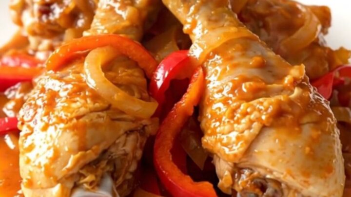 A slow cooker filled with peri peri chicken drumsticks, sliced red peppers, and onions in a vibrant red sauce.