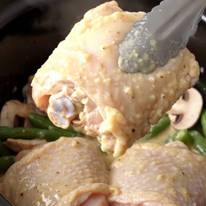 A close-up of creamy ranch mushroom chicken thighs with green beans in a black slow cooker insert.