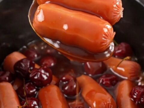 A close-up of slow cooker cocktail sausages in a thick, glossy red cherry cola glaze in a black stoneware insert.