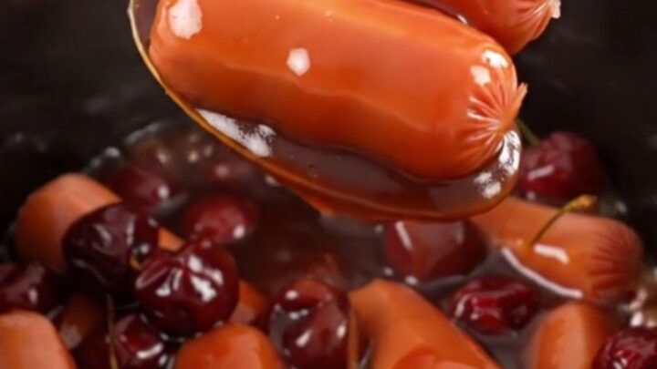 A close-up of slow cooker cocktail sausages in a thick, glossy red cherry cola glaze in a black stoneware insert.