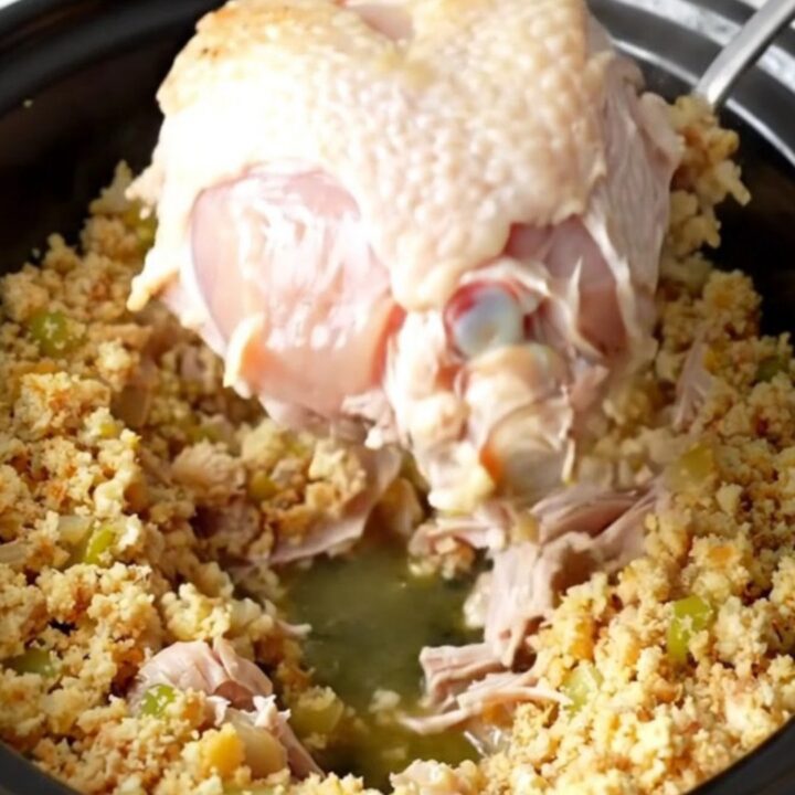 A slow cooker filled with a cooked, golden-tan turkey breast surrounded by moist, herb-flecked cornbread dressing.