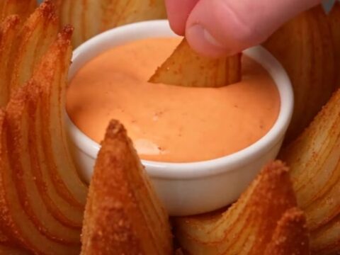 A golden-brown crispy blooming onion served on a white platter with a bowl of remoulade in the center.