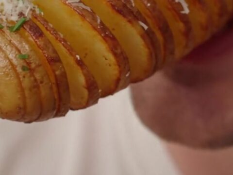 Close up of crispy golden-brown accordion potato skewers garnished with fresh chives on a wooden board.