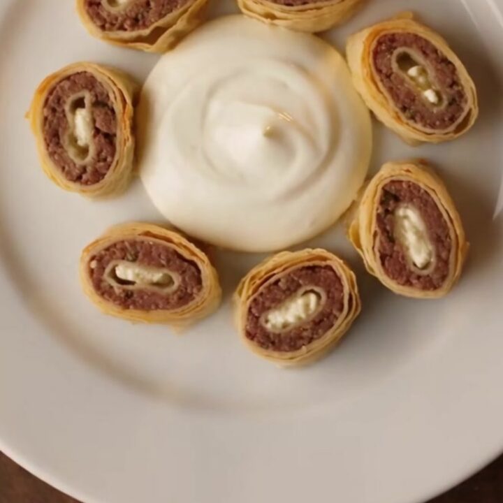 A vibrant plate of Turkish Beyti Sarma kebabs arranged in a circle with garlic yogurt and red tomato butter sauce.