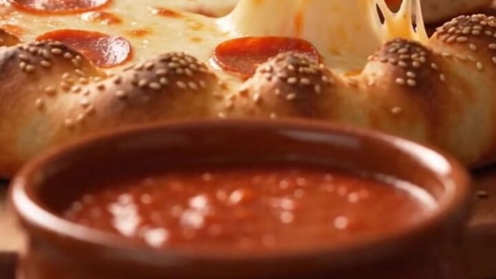 A golden-brown braided crown pizza with pepperoni and melted cheese, featuring a central bowl of marinara sauce.
