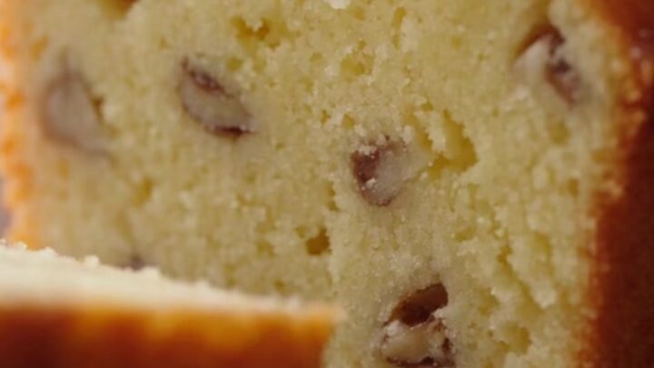 A close-up of a golden lemon loaf cake with a glistening sugar glaze and walnut flecks.