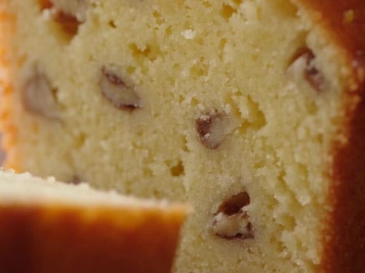 A close-up of a golden lemon loaf cake with a glistening sugar glaze and walnut flecks.