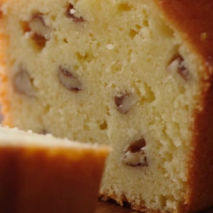 A close-up of a golden lemon loaf cake with a glistening sugar glaze and walnut flecks.