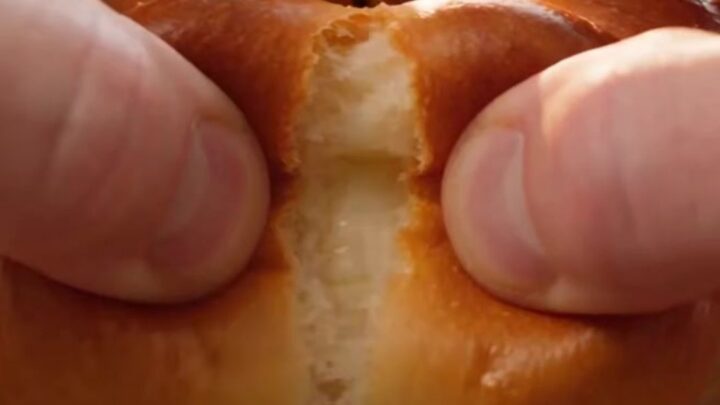 A close-up shot of a golden-brown pumpkin-shaped brioche bun being pulled apart to reveal a stretchy cheese filling.