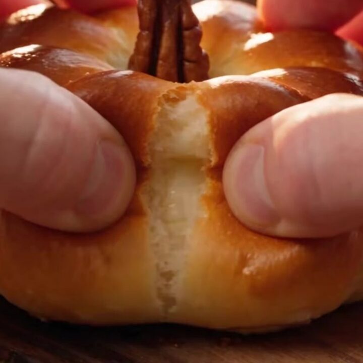 A close-up shot of a golden-brown pumpkin-shaped brioche bun being pulled apart to reveal a stretchy cheese filling.