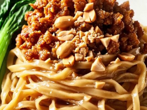 A close-up of glossy wheat noodles tossed in a rich peanut and chili oil sauce, topped with crispy pork and peanuts.