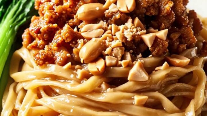 A close-up of glossy wheat noodles tossed in a rich peanut and chili oil sauce, topped with crispy pork and peanuts.
