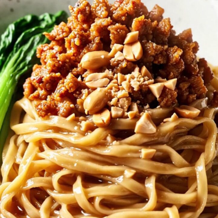 A close-up of glossy wheat noodles tossed in a rich peanut and chili oil sauce, topped with crispy pork and peanuts.