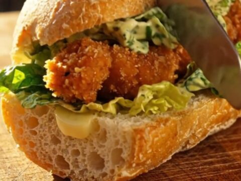A crispy chicken Caesar salad stuffed inside a warm, golden-brown garlic-buttered ciabatta baguette on a patio table.
