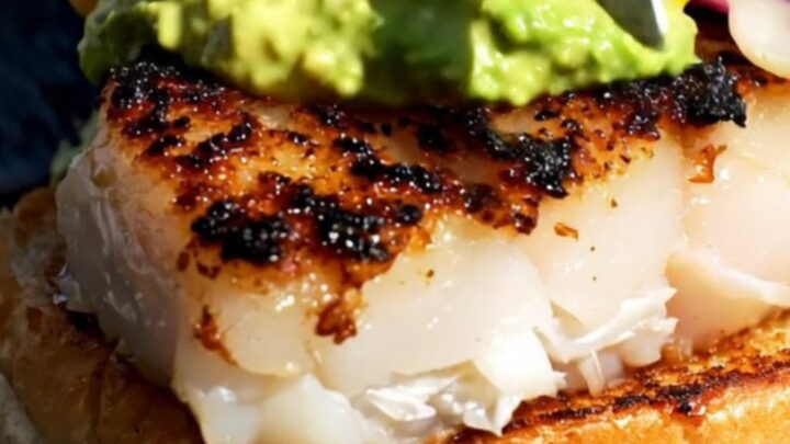 A close-up of a blackened mahi-mahi sandwich on a brioche bun with green avocado crema.