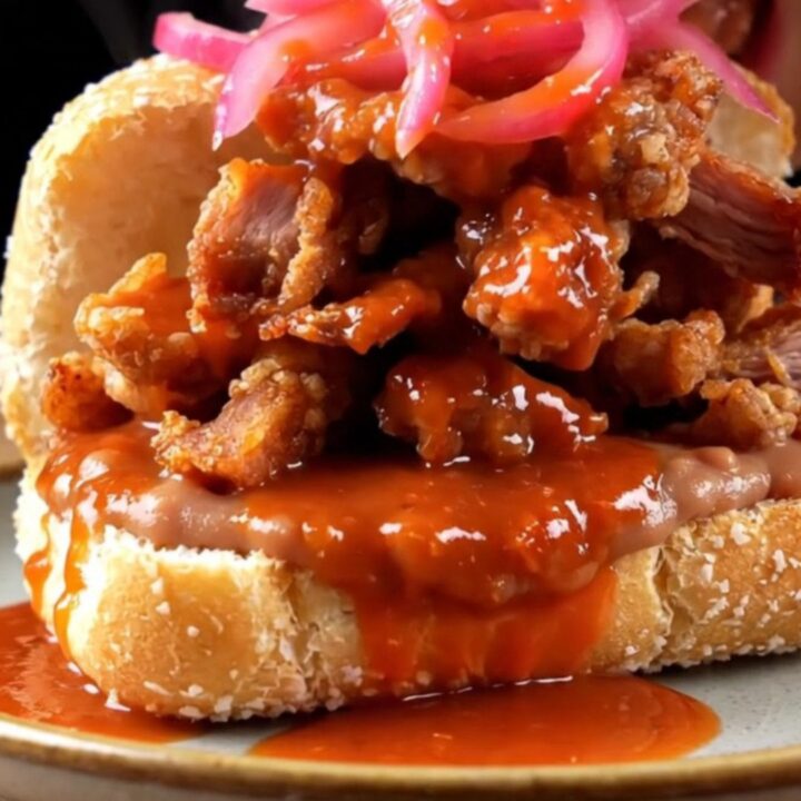 A vibrant Torta Ahogada drenched in fiery red chile sauce and topped with pickled onions on a rustic plate.