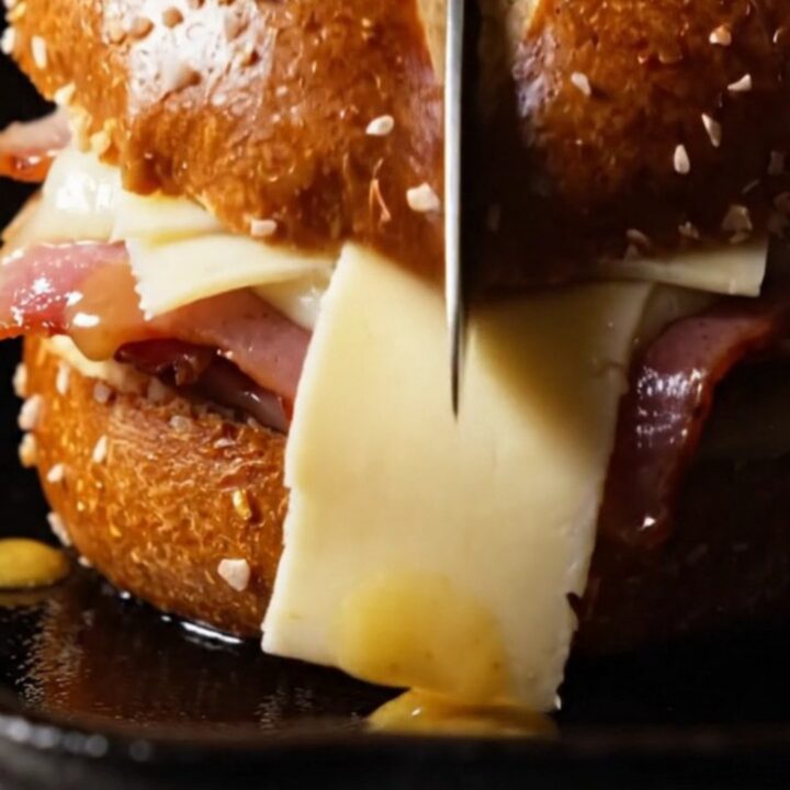 A close-up of a honey-mustard glazed ham and Gruyère sandwich on a toasted pretzel bun with visible cheese pull.