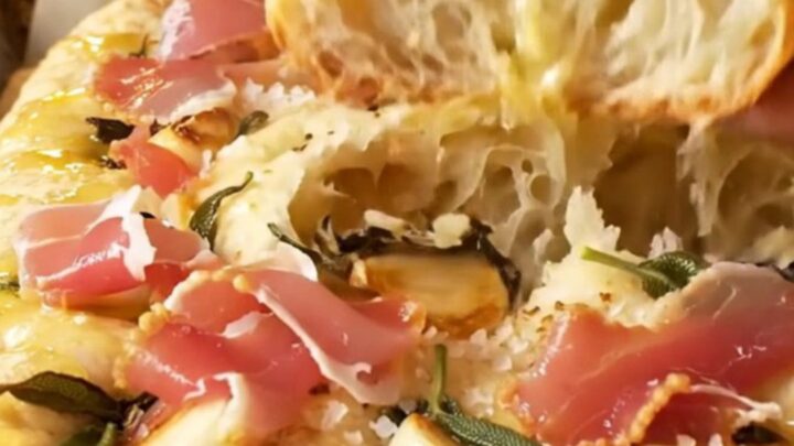 A close-up shot of a freshly baked focaccia bread with visible sage leaves, confit garlic, and prosciutto.