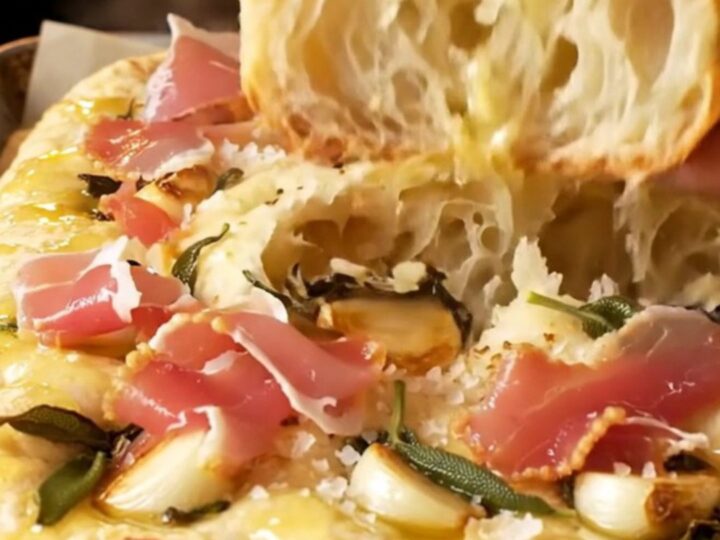A close-up shot of a freshly baked focaccia bread with visible sage leaves, confit garlic, and prosciutto.