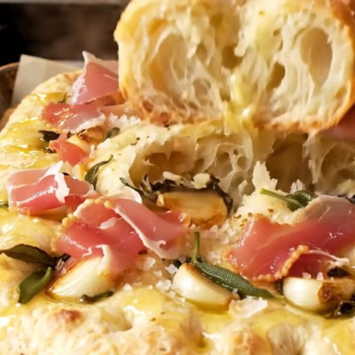 A close-up shot of a freshly baked focaccia bread with visible sage leaves, confit garlic, and prosciutto.