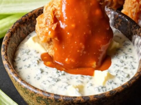 A close-up of crispy double-fried chicken wings coated in vibrant orange buffalo sauce with blue cheese crumbles.