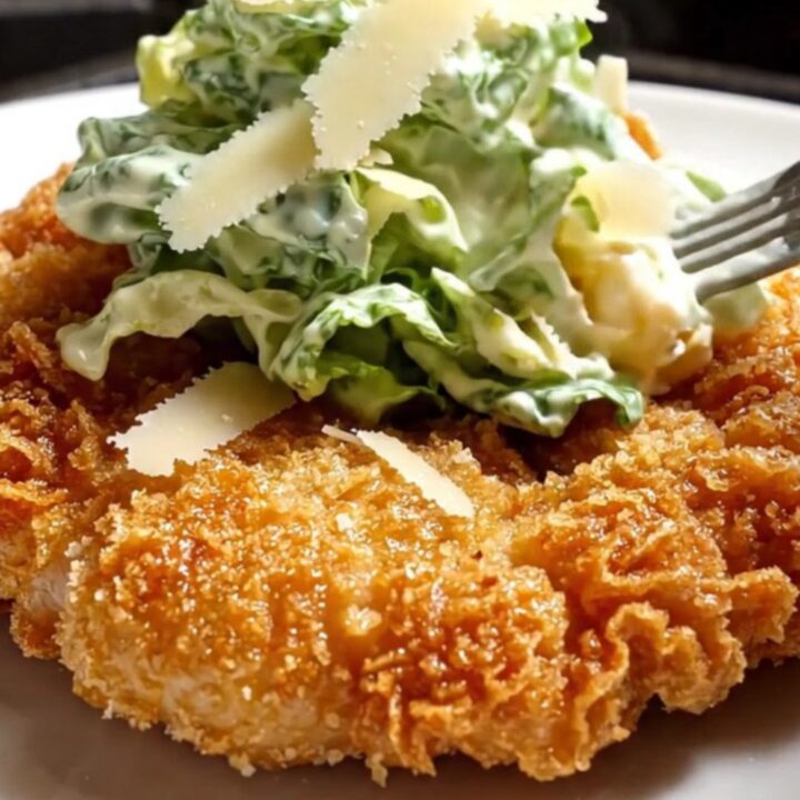 A crispy golden parmesan-crusted chicken cutlet topped with a vibrant green Caesar salad on a white plate.