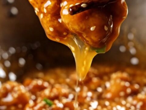 A glossy, golden-brown orange chicken piece on chopsticks with sesame seeds.