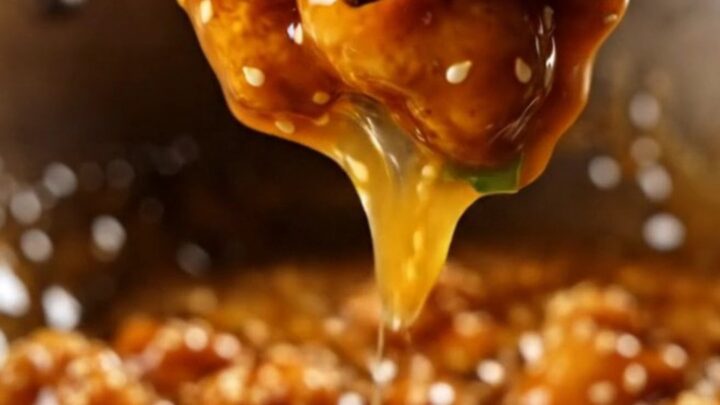 A glossy, golden-brown orange chicken piece on chopsticks with sesame seeds.