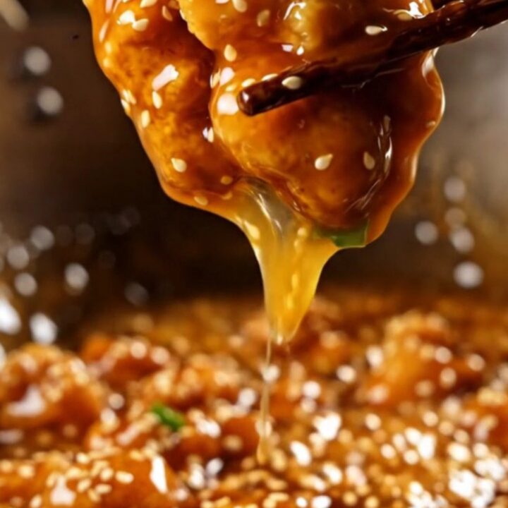 A glossy, golden-brown orange chicken piece on chopsticks with sesame seeds.