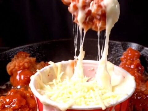 A close-up of a crispy chicken tender being lifted from a skillet, pulling long strands of melted mozzarella cheese.