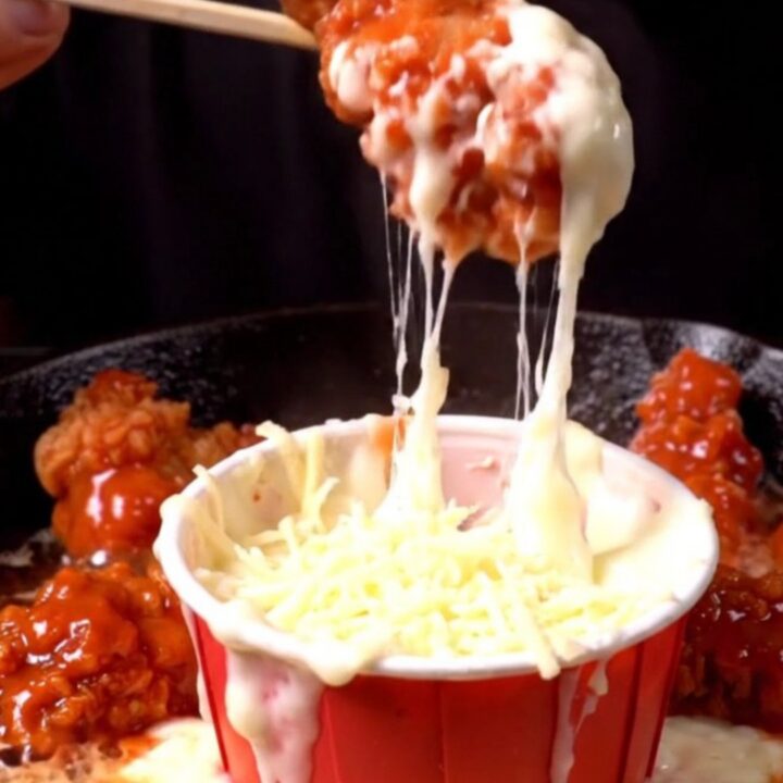 A close-up of a crispy chicken tender being lifted from a skillet, pulling long strands of melted mozzarella cheese.
