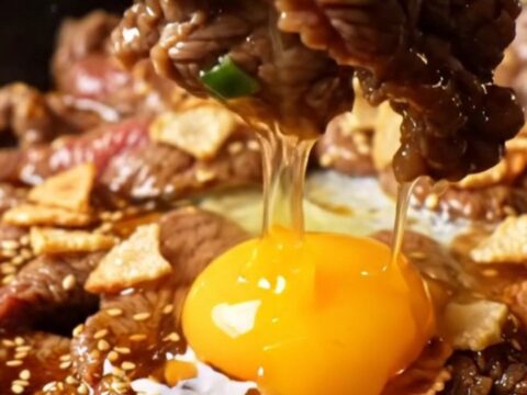 A cast-iron skillet filled with sizzling, charred ribeye bulgogi topped with golden garlic chips and a raw egg yolk.