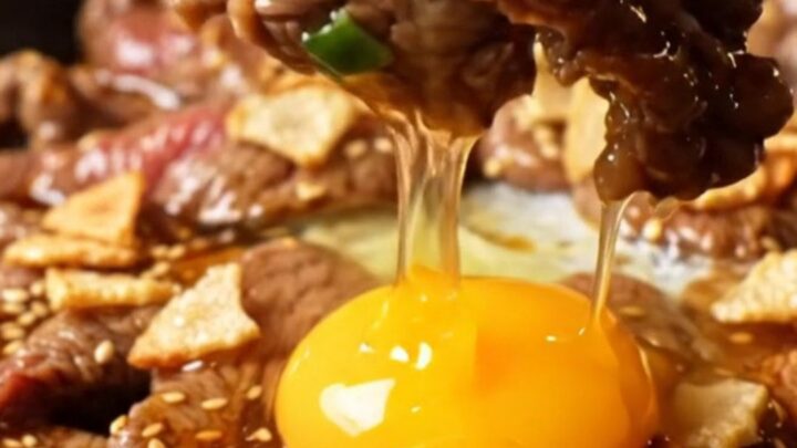 A cast-iron skillet filled with sizzling, charred ribeye bulgogi topped with golden garlic chips and a raw egg yolk.
