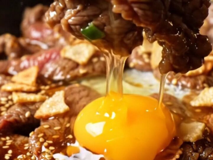 A cast-iron skillet filled with sizzling, charred ribeye bulgogi topped with golden garlic chips and a raw egg yolk.