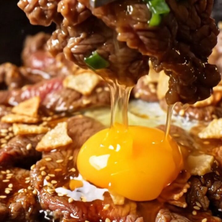 A cast-iron skillet filled with sizzling, charred ribeye bulgogi topped with golden garlic chips and a raw egg yolk.