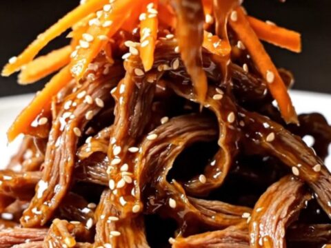 A close-up of crispy, glazed beef matchsticks piled high on a plate, garnished with vibrant orange carrots and sesame seeds.